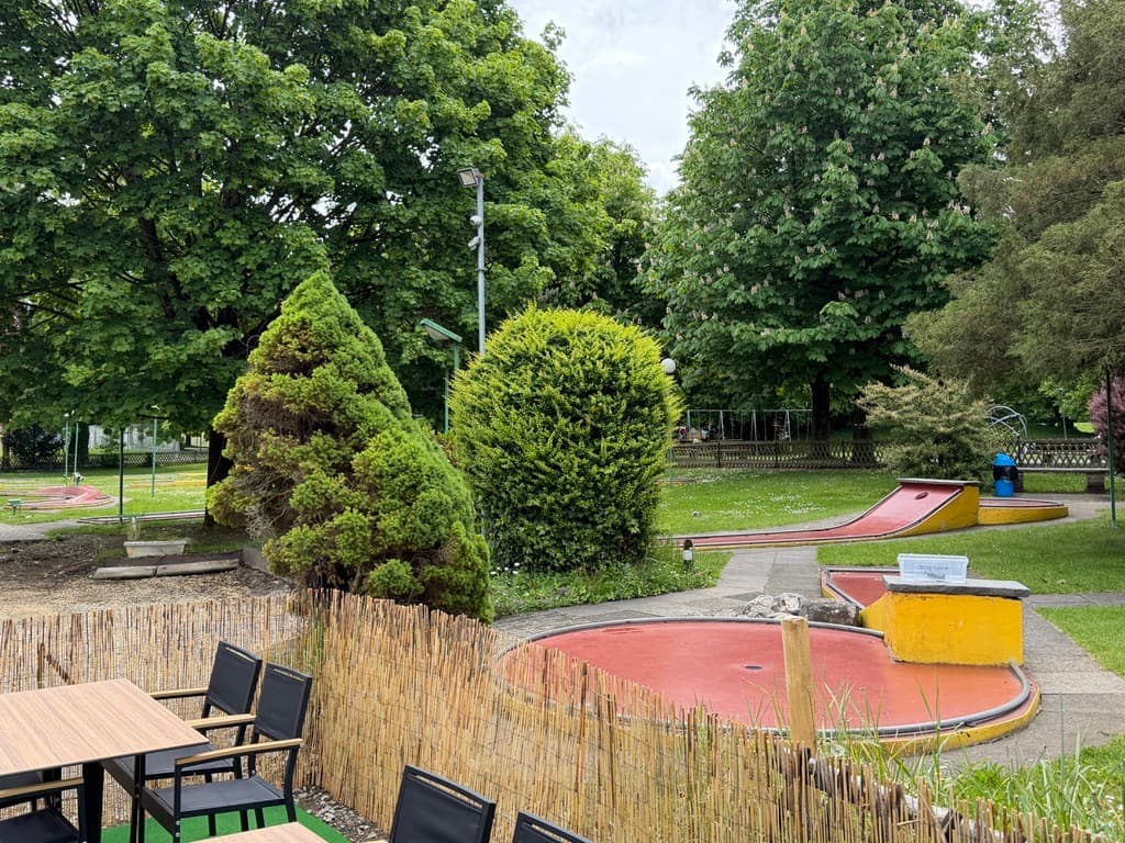 Blick von der Terrasse über die Minigolfanlage — rote und gelbe Bahnen, Hecken, Bäume.